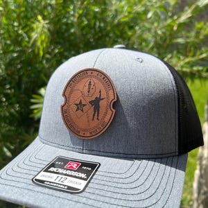 May include: A gray and black trucker hat with a brown leather patch that says "2nd Battalion 6th Marines The Ready Battalion Never To Quit". The hat is adjustable and has a Richardson logo on the side.