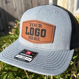 May include: A gray trucker hat with a black mesh back. The front features a brown leather patch with the text "YOUR LOGO HERE" in black. The hat has a curved brim and an adjustable strap.