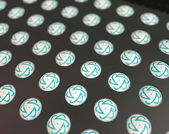 May include: A close-up shot of a black tray filled with small, round, white objects. Each object has a teal-colored, swirling design on its surface. The objects are evenly spaced and arranged in neat rows and columns, creating a visually appealing pattern.
