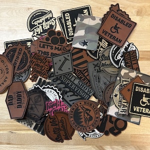 May include: A collection of leather patches in various shapes and designs. Many patches feature text, including "DISABLED VETERAN," "Salty Vet Woodworks," and "DAD in the streets, DADDY in the sheets." Colors include brown, black, and camouflage patterns.