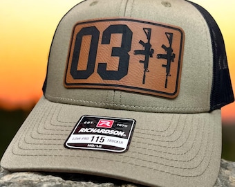 Puede incluir: Una gorra de camionero verde y negra con un parche de cuero marrón que dice "03" y tiene dos rifles estilizados. La gorra tiene una malla negra en la parte posterior y una etiqueta de Richardson en el lateral.