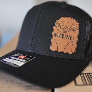 May include: A black trucker hat with a brown leather patch featuring a box of crayons with the word "MARINE" written on it.