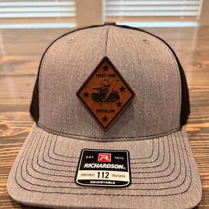 May include: A gray and black trucker hat with a brown leather patch that says "First Tank Battalion" with a star and a tank graphic. The hat has a black mesh back and a black brim. The hat also has a black and red label that says "Est. 1970 Richardson Original 112 Trucker Adjustable".