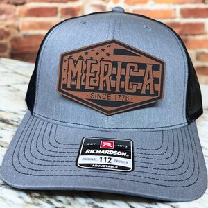 May include: A gray and black trucker hat with a brown leather patch that says "MERICA SINCE 1776" with a small American flag design. The hat is adjustable and has a Richardson 112 label.