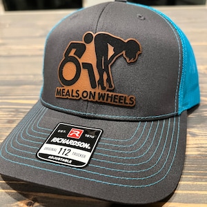 May include: A black and turquoise trucker hat with a brown leather patch that says "Meals on Wheels" and a graphic of a person riding a motorcycle. The hat is adjustable and has a Richardson 112 label.