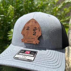 May include: A gray and black trucker hat with a brown leather patch featuring a sword and the text "2 2 Warlords Afghanistan Iraq Kuwait Lebanon Haiti Cuba Taraawa". The hat also has a black and red label with the text "EST. 1870 RICHARDSON ORIGINAL 112 TRUCKER ADJUSTABLE".
