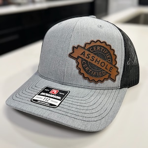 May include: A gray and black trucker hat with a brown leather patch that says "Certified Asshole Certified". The hat is adjustable and has a Richardson 112 label.