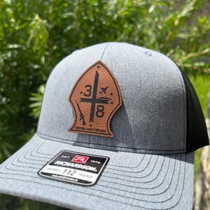 May include: A gray and black trucker hat with a brown leather patch featuring a sword, a number 3, and the text "FORTUNA FAVET FORTIBUS". The hat has a black mesh back and a Richardson 112 adjustable buckle.