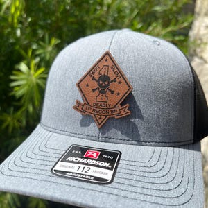 May include: A gray and black trucker hat with a brown leather patch featuring a skull and crossbones with the text "Swift, Silent, Deadly, 1st Recon Bn". The hat has a Richardson 112 label.
