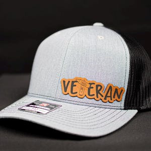May include: A gray and black trucker hat with a brown leather patch that says "Veteran" with a small wrench graphic.
