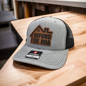 May include: A gray and black trucker hat with a brown leather patch that says "Defund the HOA". The hat has a black mesh back and a Richardson 112 adjustable label.