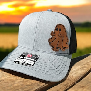 May include: A gray and black trucker hat with a brown leather patch of a ghost giving the middle finger. The hat has a black mesh back and a gray front with the text "Richardson 112" on the front.