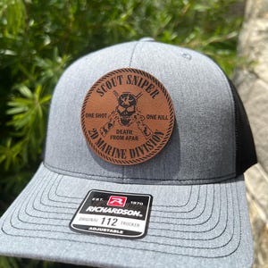 May include: A gray and black trucker hat with a brown leather patch that reads "Scout Sniper One Shot Death From Afar One Kill 2D Marine Division". The hat is adjustable and has a Richardson logo.