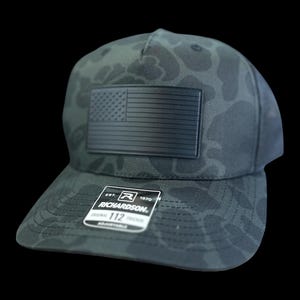 May include: A black camouflage Richardson 112 trucker hat with a black American flag patch. The hat has a curved brim and an adjustable snapback closure. The tag reads "Richardson 112 Trucker".