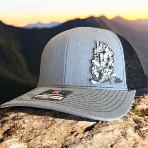 May include: A gray trucker hat with a black mesh back and a curved brim. The hat features a black and white graphic of a muscular boar holding a weapon, with skulls at its feet. The hat has an adjustable strap.
