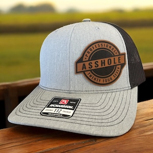 May include: Gray and black trucker hat with a leather patch that reads "PROFESSIONAL ASSHOLE TWENTY FOUR SEVEN". The hat has a curved brim and a mesh back. The Richardson brand label is visible.