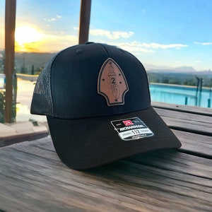 May include: Black and gray mesh trucker hat with a brown leather patch featuring a torch and the number "2" surrounded by stars. The patch is shaped like a spearhead. The hat has an adjustable strap with the text "Richardson 112 Original Trucker Adjustable" printed on it.