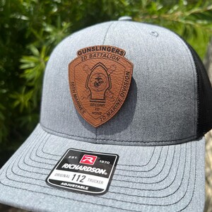 May include: A gray and black trucker hat with a brown leather patch that says "GUNSLINGERS 2D BATTALION SECOND TO NONE 2D MARINE DIVISION 10TH MARINES". The hat also has a red "R" logo with the text "EST. 1970 RICHARDSON® ORIGINAL 112 TRUCKER ADJUSTABLE".