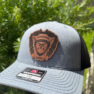 May include: A gray and black trucker hat with a brown leather patch featuring a bear head, crossed swords, and the text "THUNDERING THIRD BATTALION", "EST. 1870", and "RICHARDSON 112".