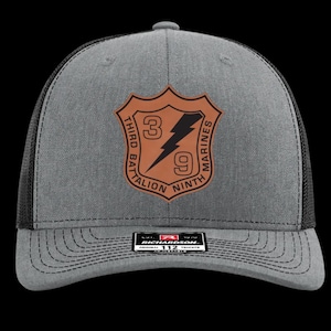 May include: Gray trucker hat with a black mesh back. The front features a brown leather patch with the text "THIRD BATTALION NINTH MARINES" around a shield with the numbers "3" and "9" and a lightning bolt. The hat has a curved brim.