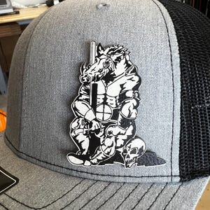 以下が含まれることがあります： グレーと黒のトラッカーハット。イノシシの戦士が槍を持ち、足元に2つのドクロがある白黒グラフィック。ハットはメッシュバックとカーブしたつばが付いています。