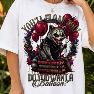 Könnte beinhalten: Ein weißes T-Shirt mit einer Grafik eines als Clown verkleideten Waschbären, der auf einem Stapel Bücher sitzt. Der Waschbär hält rote Luftballons. Der Text lautet: YOU'LL FLOAT TOO! DO YOU WANT A BALLOON? Mit lila Rosen und Kürbissen.