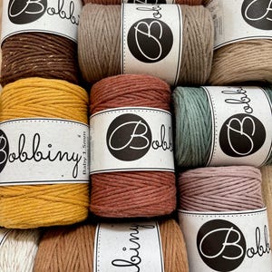 Könnte beinhalten: Eine Sammlung von zehn Spulen Bobbiny Baby 1,5 mm Makramee-Garn in verschiedenen Farben wie Senfgelb, Terrakotta, Salbeigrün, Altrosa, Braun mit Goldakzenten und Beige, jeweils mit einem weißen Papieretikett mit dem Markennamen versehen.