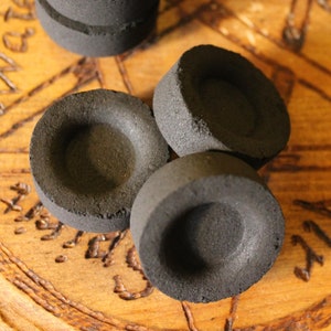 Könnte beinhalten: Vier schwarze Holzkohle-Scheiben mit einem kleinen Loch in der Mitte, gestapelt auf einer Holzoberfläche mit einem geschnitzten Design.
