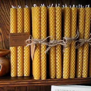 Può includere: Una collezione di candele in cera d'api giallo dorato con una trama a nido d'ape, legate con spago. Un piccolo diffusore di oli essenziali in ceramica con un ritaglio a forma di mezzaluna è posto su una mensola di legno. Un'etichetta è attaccata alle candele.