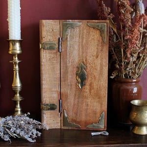 May include: A rectangular wooden box with a natural light brown finish, featuring brass corner accents, two brass hinges on the left side, and a central brass decorative plate. A small metal key lies on the surface in front of the wooden box.