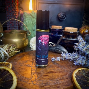 Puede incluir: Un pequeño frasco de vidrio con un aplicador de bola negro. El frasco está etiquetado con "Empress of the Moon" y "Fast Luck" con las palabras "Patchouli - Rose - Mint" debajo. El frasco está sentado sobre una superficie de madera con un diseño circular.