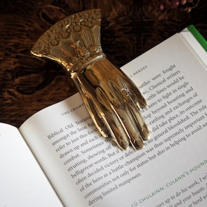Könnte beinhalten: Ein goldfarbener, handförmiger Lesezeichen liegt auf den Seiten eines Buches. Das Lesezeichen hat aufwendige Details und besteht aus glänzendem Metall. Die Buchseiten sind weiß mit schwarzem Text, die Kanten sind grün.