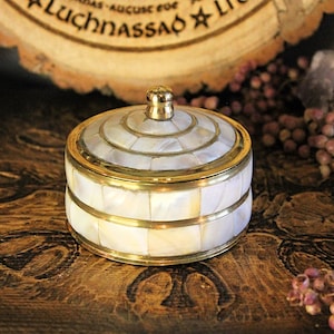 Könnte beinhalten: Eine runde Schmuckdose mit Deckel, verziert mit einem Perlmutt-Mosaik-Design und Messingbesatz. Die Dose hat einen goldfarbenen Knopf oben. Dekorationsobjekt auf dunkler Holzoberfläche.
