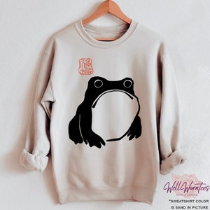 Puede incluir: Una sudadera beige con una ilustración de rana negra y un sello rojo con un carácter chino. La sudadera está en una percha.