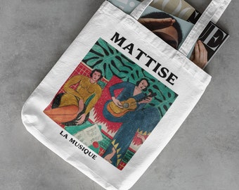 Puede incluir: Bolso tote blanco con una impresión de la obra "La Musique" de Matisse. El bolso muestra la palabra "MATISSE" en negro sobre la obra de arte y "LA MUSIQUE" debajo. Se ven revistas dentro del bolso.
