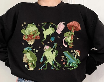 Puede incluir: Sudadera negra con un estampado gráfico de ranas verdes en varias poses, algunas sentadas en hongos, otras bailando. Las ranas están rodeadas de pequeñas flores blancas y puntos negros.