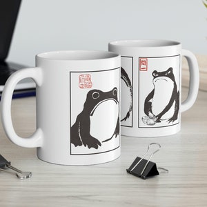 May include: Two white ceramic mugs with black and white illustrations of frogs. The mugs have a black outline and a red stamp with Japanese characters. One frog is sitting and the other is standing.