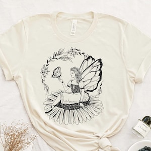 May include: A white t-shirt featuring a black and white illustration of a fairy sitting on a flower with a butterfly. The fairy has wings and is holding a butterfly in her hand.