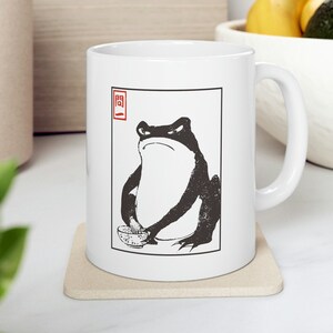 May include: White ceramic mug featuring a black and white illustration of a frog holding a small bowl. The frog has an angry expression. The mug has a handle and a red and black Japanese-style design.