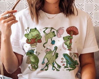 Pode incluir: Camiseta branca com uma estampa gráfica de sapos verdes e azuis em cogumelos. Os sapos estão em várias poses, incluindo sentados, pulando e segurando guarda-chuvas.