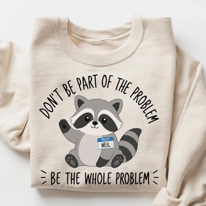 Peut inclure: Un sweat-shirt à col rond couleur crème avec une illustration de raton laveur portant un badge bleu indiquant NEIL. Le texte au-dessus du raton laveur dit DON'T BE PART OF THE PROBLEM et le texte en dessous dit BE THE WHOLE PROBLEM.