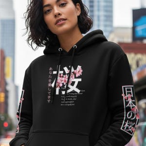 May include: Black hoodie with a front graphic featuring Japanese characters and pink cherry blossoms. The sleeves also have Japanese text and floral accents. The hoodie has a drawstring hood and a front pocket.
