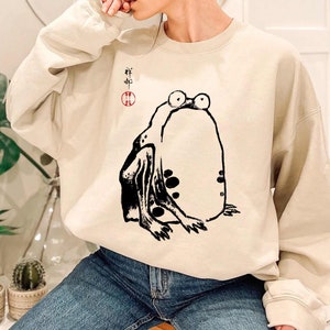 May include: A beige sweatshirt with a black and white illustration of a frog. The frog is sitting on its hind legs with its front legs outstretched. The sweatshirt has a red circle with Japanese characters in it.
