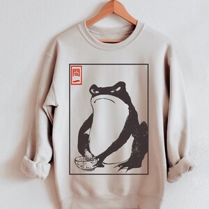 May include: A beige sweatshirt with a black and white illustration of a frog holding a bowl. The frog is sitting in a square frame with the Japanese character "開" in red in the top left corner.