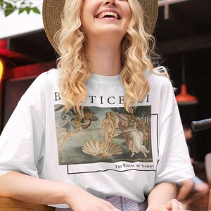 May include: A white t-shirt with a print of the painting "The Birth of Venus" by Sandro Botticelli. The shirt has a crew neck and short sleeves. The model is wearing a straw hat and smiling.