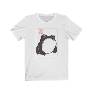 May include: White t-shirt with a black and white illustration of a frog in a square frame. The frog is looking up and has a large, round belly. The word "蛙" is written in red in the top left corner of the frame.