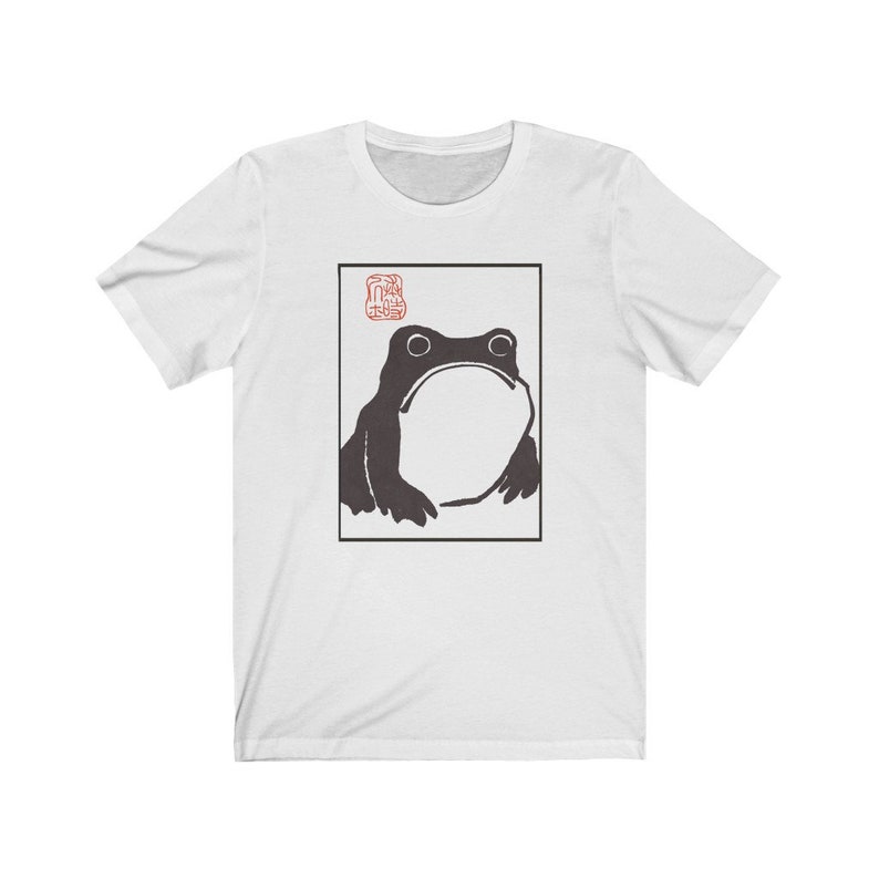 May include: White t-shirt with a black and white illustration of a frog in a square frame. The frog is looking up and has a large, round belly. The word "蛙" is written in red in the top left corner of the frame.
