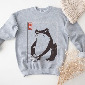 May include: A gray sweatshirt with a black and white illustration of a frog holding a bowl. The illustration is in a white frame with the word "関" in red in the top left corner.