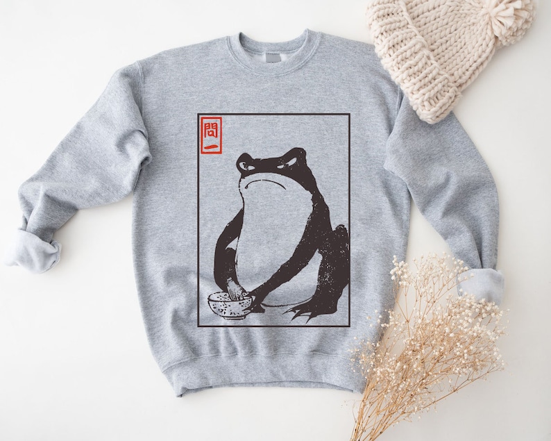 May include: A gray sweatshirt with a black and white illustration of a frog holding a bowl. The illustration is in a white frame with the word "関" in red in the top left corner.