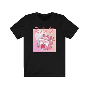 Op de afbeelding: Zwart T-shirt met een afbeelding van een roze aardbeienmelkpak en Japanse tekst. Het ontwerp bevat een roze en blauw raster, een witte cirkel en het woord "MILK" in roze.